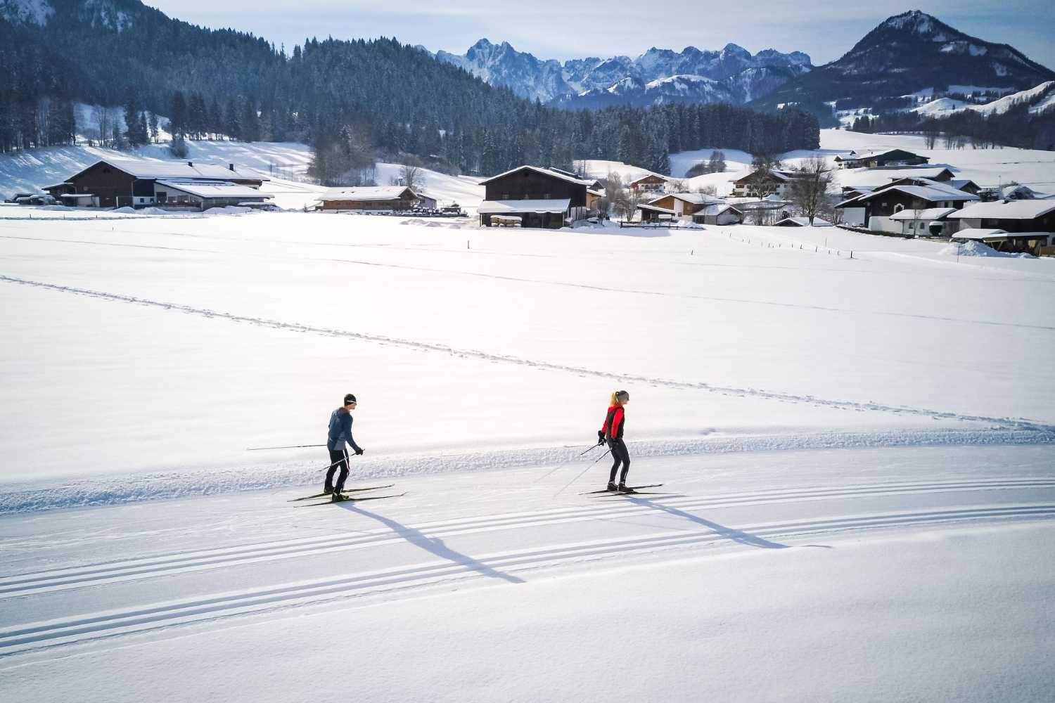 Zwei Langläufer auf Loipe vor verschneitem Dorf und Bergkulisse im Kaiserwinkl in Tirol