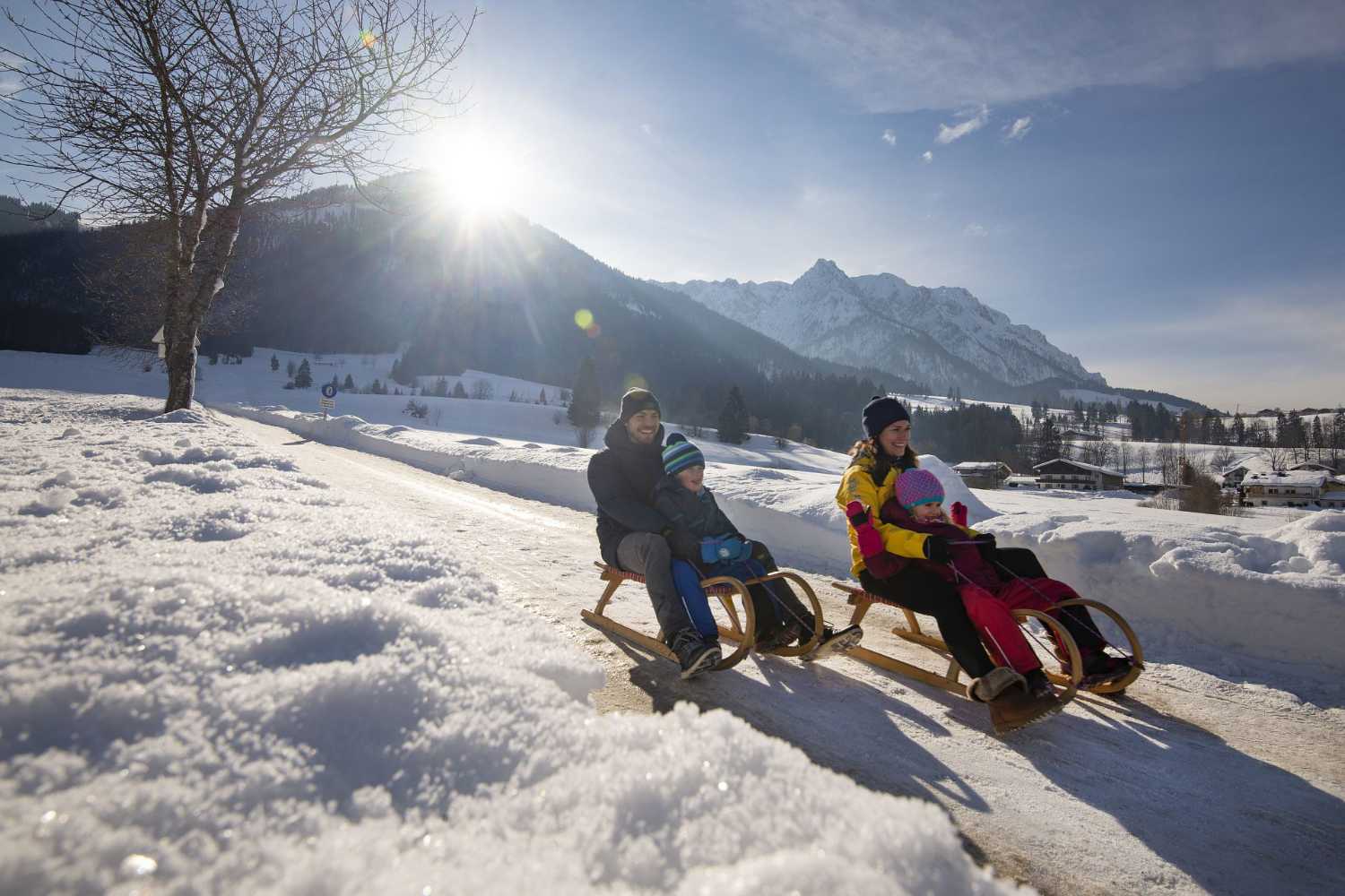 Familie beim Rodeln auf verschneitem Weg im Kaiserwinkl in Tirol mit Wintersonne und Bergblick