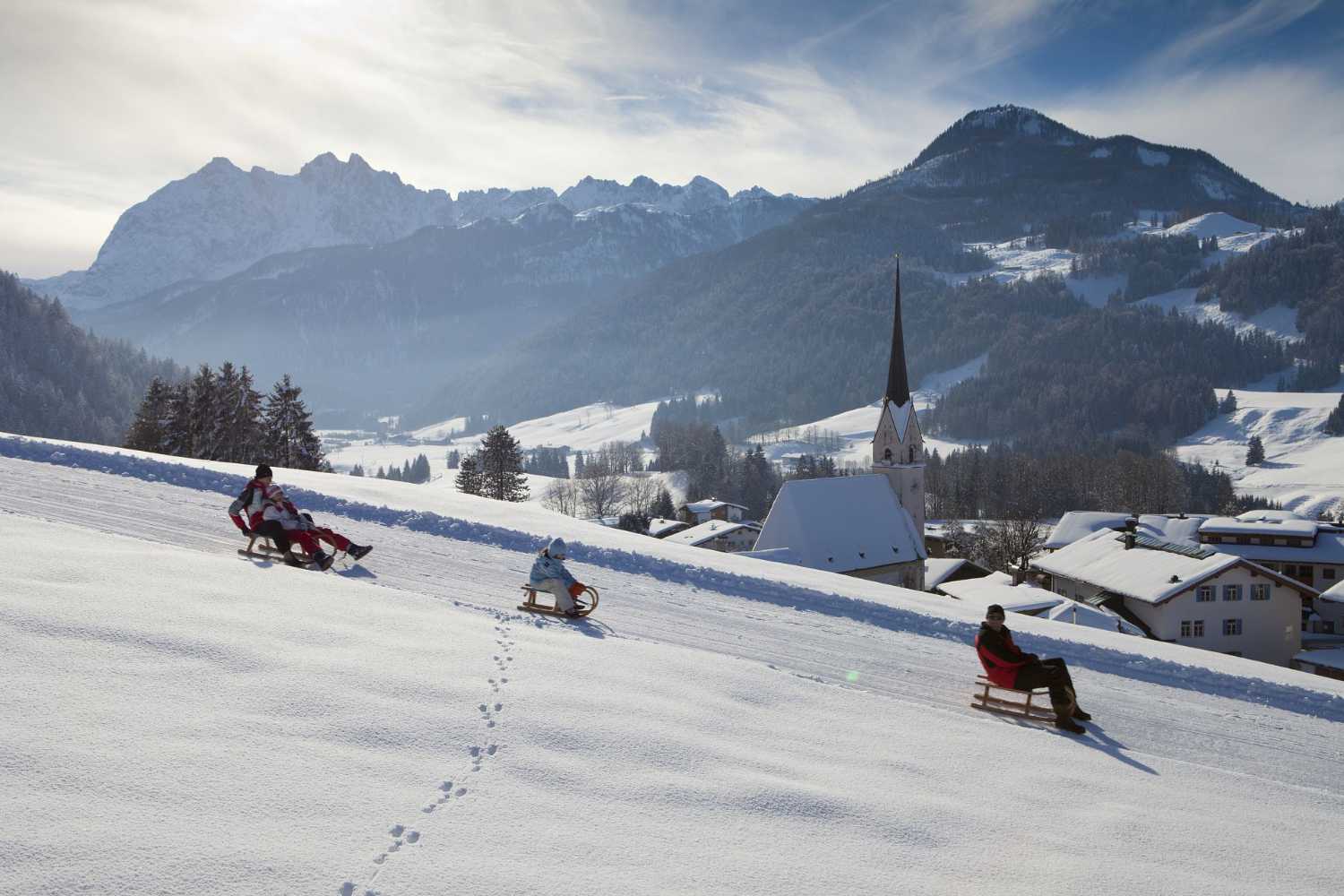 Rodeln am Hang mit Blick auf ein verschneites Dorf und Kirche im Kaiserwinkl in Tirol