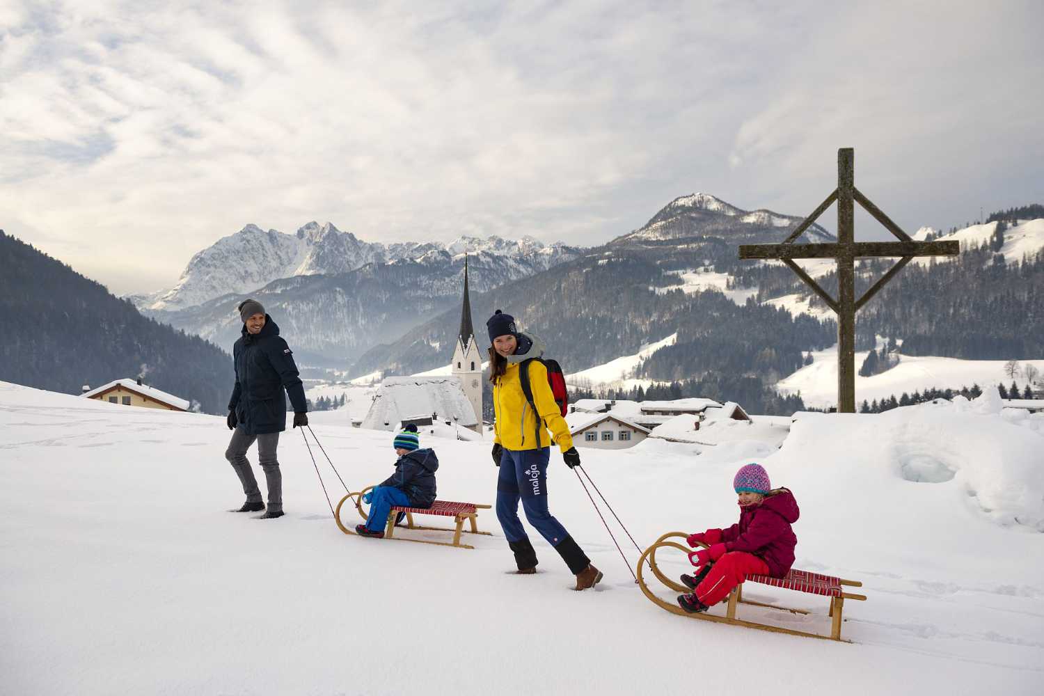 Familie mit Schlitten im Schnee nahe Dorfkirche und Bergpanorama im Kaiserwinkl in Tirol